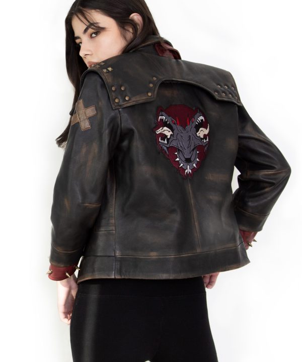 Photo of a model wearing a dark brown leather jacket. The back of the jacket is visible, showing the same 3/4 length red studded leather cuffs and the light brown X on the left upper arm. The back of the jacket has a large patch, showing a two-headed dog wearing a studded collar, both heads with their mouths open and teeth bared. The background of the patch is in the shape of an upside-down teardrop.The jacket pictured is the Vi arcane season 2 jacket.